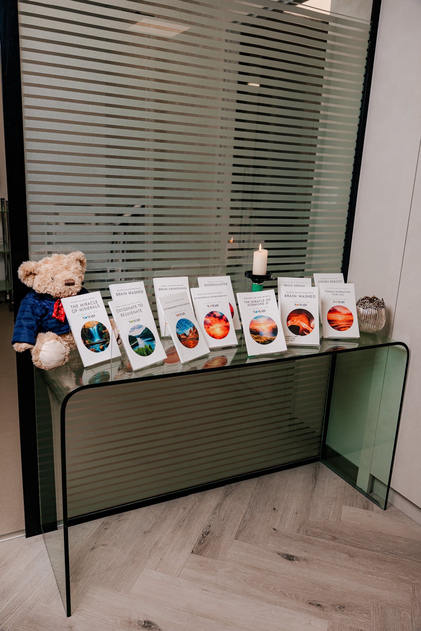Glass console table in a Levitas clinic displaying a row of NOMAD books, a lit candle and a teddy bear in a blue jacket